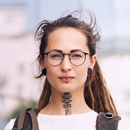 Mujer joven con gafas y dreadlocks mostrando tatuaje de rosa en cuello estilo boho