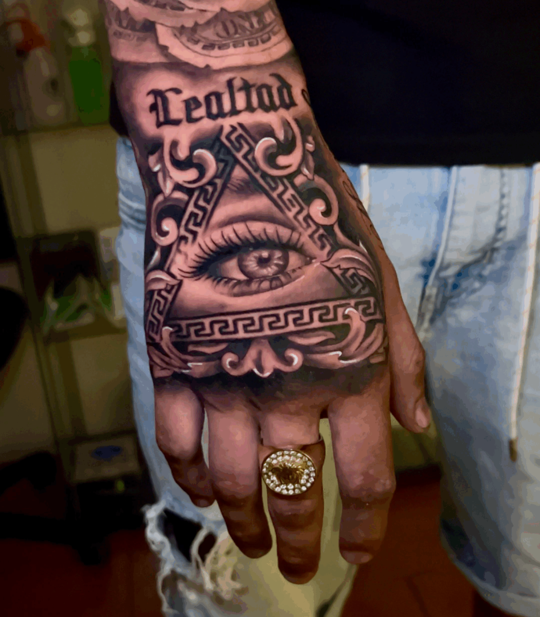 Tatuaje en mano con ojo, texto Realidad y elementos decorativos chicanos en estilo realista